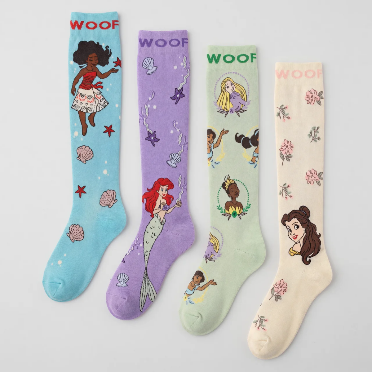 Flat lay display featuring four pairs of Disney Princess knee-high compression socks lined up side-by-side, including Moana (aqua), Ariel (purple), Multi-Princess (mint green), and Belle (cream), all featuring "WOOF" top cuff branding.