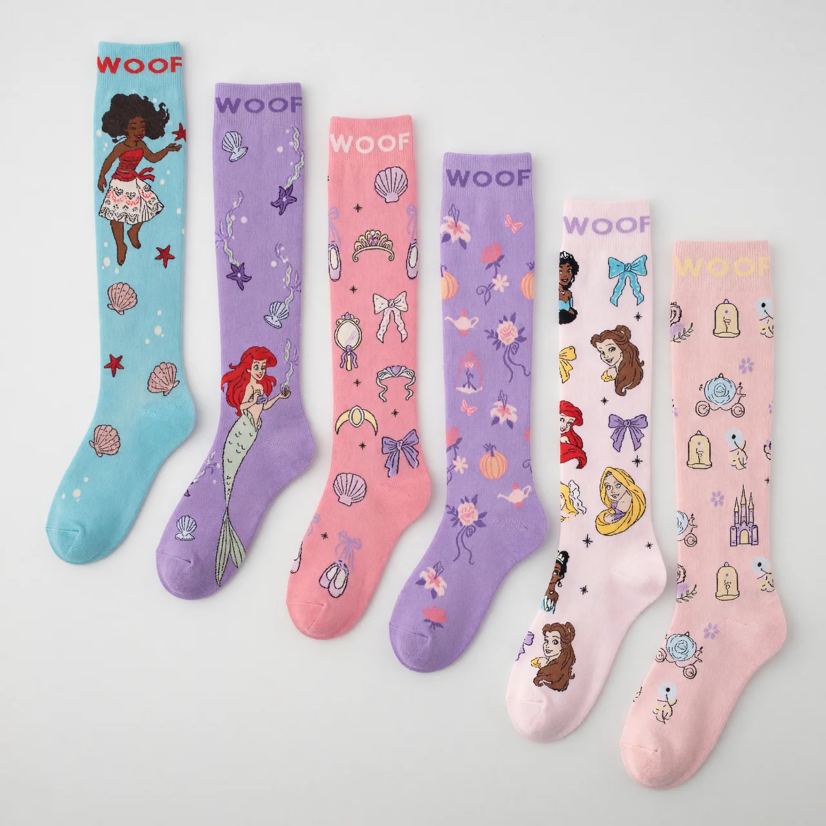 Flat lay display showcasing seven pairs of Disney Princess knee-high compression socks lined up side-by-side, including Moana (aqua), Ariel (purple), Princess Icons (pink), Magic Carriage & Lamp (lilac), Character Collage (light pink), and Cinderella (soft pink), all featuring "WOOF" top cuff branding.